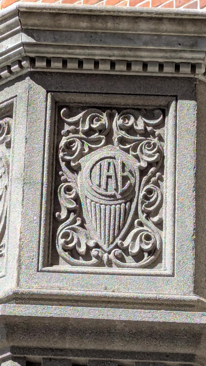 Un detalle magnífico. Es el bajorrelieve tallado en piedra que viste en la foto anterior, ahora de cerca. Se puede apreciar claramente el monograma CIAE dentro de un círculo, rodeado de volutas y hojas de estilo art nouveau, todo sobre lo que parece ser una lira o instrumento de cuerdas estilizado en la parte inferior. Es un trabajo de cantería muy fino, típico de la arquitectura industrial de principios del siglo XX con aspiraciones estéticas elevadas. Un detalle que hoy poca gente nota al pasar.