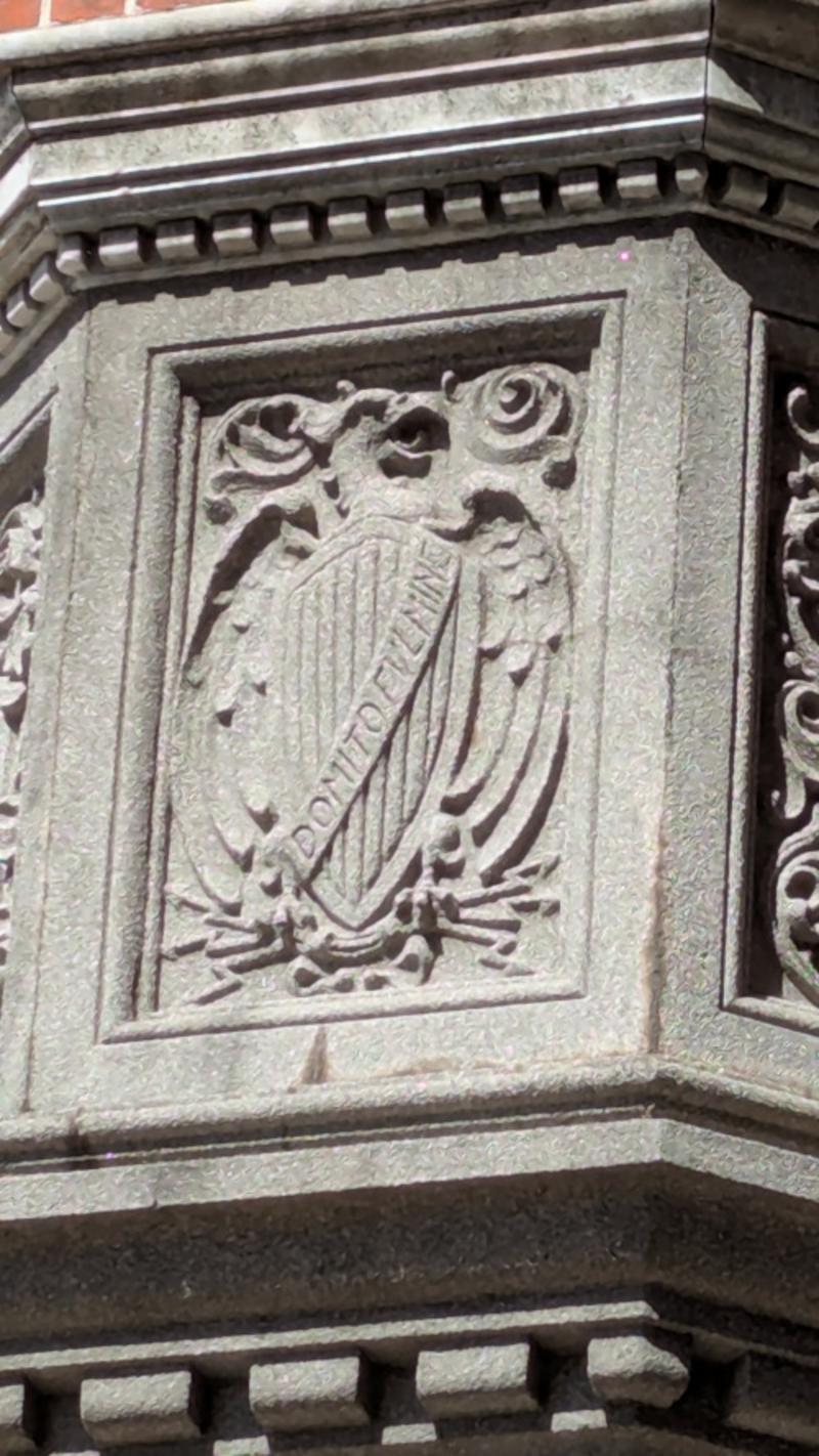 Bajorrelieve tallado en piedra gris sobre uno de los paneles del portal de entrada de la Usina del Arte, Buenos Aires. Representa un escudo heráldico con un águila de alas desplegadas encima, rodeado de volutas decorativas en la parte superior. Atravesando el escudo en diagonal, una filacteria lleva inscrita la frase en latín DOMITO FVLMINE, que significa 'domado el rayo', lema de la Compañía Alemana de Electricidad que construyó el edificio en 1916. El conjunto simboliza el dominio humano sobre la electricidad.
