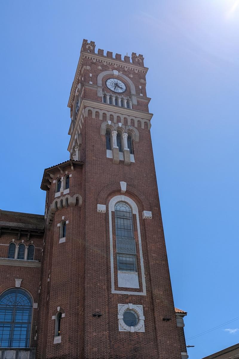 Torre de ladrillo rojo de la Usina del Arte, fotografiada en contrapicado desde la calle contra un cielo azul despejado. La torre se eleva en varios cuerpos: en la base, una ventana alta de arco apuntado con marco de piedra clara; en el cuerpo medio, un grupo de tres ventanas geminadas con columnitas; cerca de la cúspide, un reloj circular embutido en un arco de medio punto; y en lo más alto, una corona de almenas de estilo medievalizante. Los detalles ornamentales en piedra blanca contrastan con el ladrillo rojizo. A la izquierda se ve parte del cuerpo principal del edificio con ventanas de arco semicircular. El estilo evoca las torres cívicas del norte de Italia, aunque el edificio fue construido como usina eléctrica en Buenos Aires en 1916.